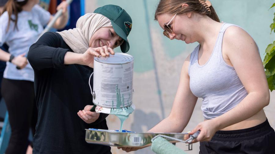 Two students work together to pour the primer paint from a bucket into a tray.